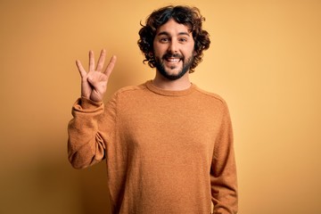 Young handsome man with beard wearing casual sweater standing over yellow background showing and pointing up with fingers number four while smiling confident and happy.