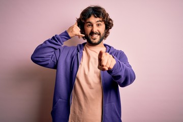 Young handsome sporty man with beard wearing casual sweatshirt over pink background smiling doing talking on the telephone gesture and pointing to you. Call me.