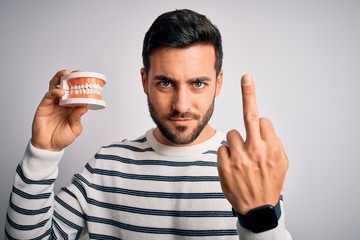 Young handsome man with beard holding plastic denture teeth over white background Showing middle...
