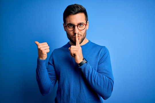 Young handsome man with beard wearing casual sweater and glasses over blue background asking to be quiet with finger on lips pointing with hand to the side. Silence and secret concept.