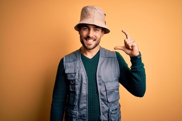 Handsome tourist man with beard on vacation wearing explorer hat over yellow background smiling and confident gesturing with hand doing small size sign with fingers looking and the camera. Measure