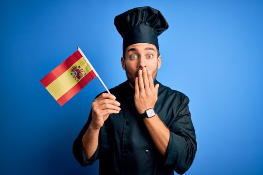Young handsome cooker man with beard wearing uniform holding spain spanish flag cover mouth with hand shocked with shame for mistake, expression of fear, scared in silence, secret concept