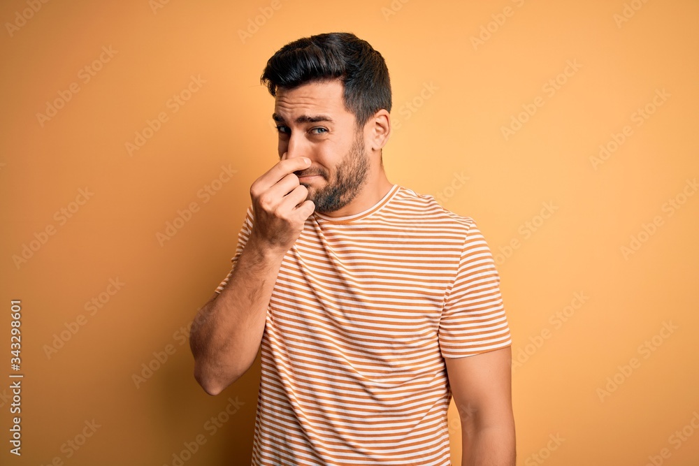 Canvas Prints Young handsome man with beard wearing casual striped t-shirt over yellow background smelling something stinky and disgusting, intolerable smell, holding breath with fingers on nose. Bad smell