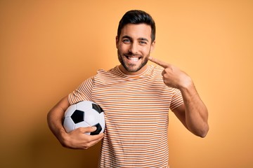 Handsome player man with beard playing soccer holding footballl ball over yellow background smiling cheerful showing and pointing with fingers teeth and mouth. Dental health concept.