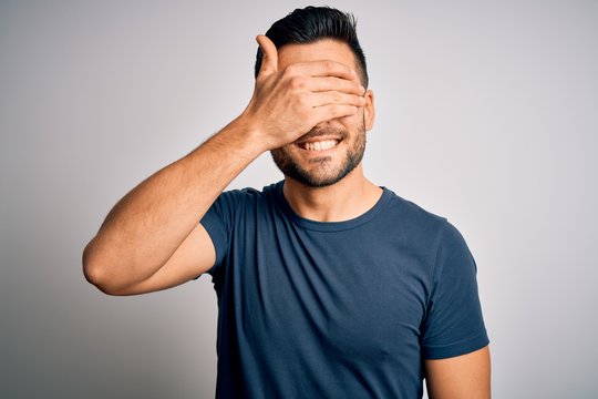Young handsome man wearing casual t-shirt standing over isolated white background smiling and laughing with hand on face covering eyes for surprise. Blind concept.