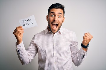 Young business man holding fake news paper over isolated background screaming proud and celebrating...