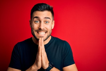 Fototapeta na wymiar Young handsome man wearing casual black t-shirt standing over isolated red background praying with hands together asking for forgiveness smiling confident.