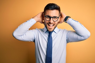 Young handsome businessman wearing tie and glasses standing over yellow background relaxing and stretching, arms and hands behind head and neck smiling happy