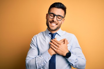 Young handsome businessman wearing tie and glasses standing over yellow background smiling with hands on chest with closed eyes and grateful gesture on face. Health concept.