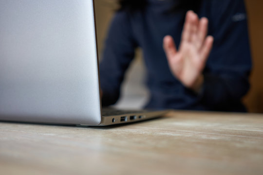 Young woman working from home uses a laptop computer at home to join a video conference meeting and waving hand to her colleagues. Selective focus on the edge of the laptop.