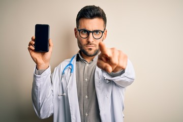 Young doctor man wearing stethoscope showing smartphone screen over isolated background pointing with finger to the camera and to you, hand sign, positive and confident gesture from the front