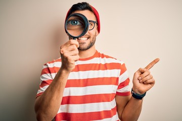 Young detective man looking through magnifying glass over isolated background very happy pointing...