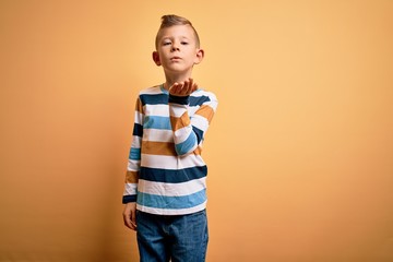 Young little caucasian kid with blue eyes wearing colorful striped shirt over yellow background looking at the camera blowing a kiss with hand on air being lovely and sexy. Love expression.