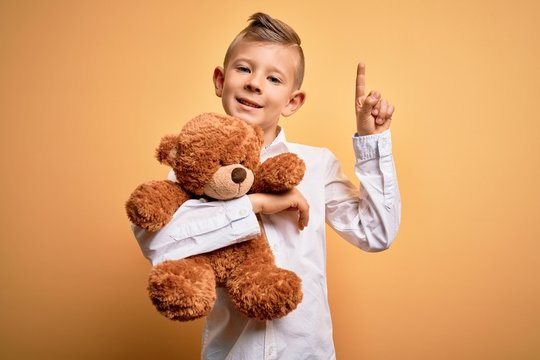 Young little caucasian kid hugging teddy bear stuffed animal over yellow background surprised with an idea or question pointing finger with happy face, number one