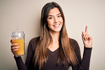 Young beautiful girl drinking glass of healthy orange juice over isolated white background surprised with an idea or question pointing finger with happy face, number one