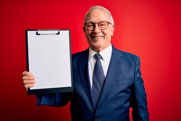 Senior grey haired business man holding clipboard over red background with a happy face standing...