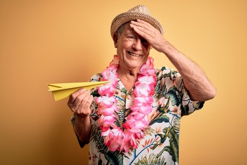 Grey haired senior man wearing summer hat and hawaiian lei holding paper plane on vacation stressed with hand on head, shocked with shame and surprise face, angry and frustrated. Fear and upset