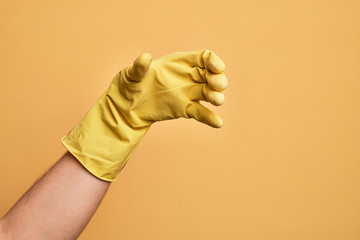 Hand of caucasian young man with cleaning glove over isolated yellow background holding invisible object, empty hand doing clipping and grabbing gesture