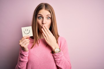 Fototapeta premium Young beautiful redhead woman holding reminder paper with smile emoji message cover mouth with hand shocked with shame for mistake, expression of fear, scared in silence, secret concept