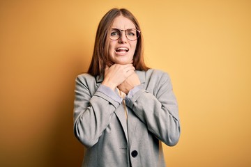 Young beautiful redhead woman wearing jacket and glasses over isolated yellow background shouting and suffocate because painful strangle. Health problem. Asphyxiate and suicide concept.