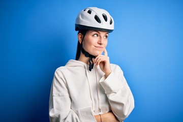 Young beautiful redhead cyclist woman wearing bike helmet over isolated blue background with hand on chin thinking about question, pensive expression. Smiling with thoughtful face. Doubt concept.