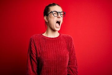 Young beautiful redhead woman wearing casual sweater over isolated red background angry and mad screaming frustrated and furious, shouting with anger. Rage and aggressive concept.