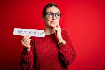 Young beautiful redhead woman holding paper with love yourself message serious face thinking about...