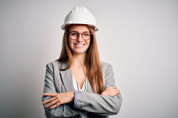 Young beautiful redhead architect woman wearing security helmet over white background happy face smiling with crossed arms looking at the camera. Positive person.
