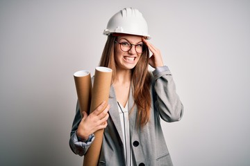 Young beautiful redhead architect woman wearing glasses and safety helmet holding blueprints stressed with hand on head, shocked with shame and surprise face, angry and frustrated. Fear and upset