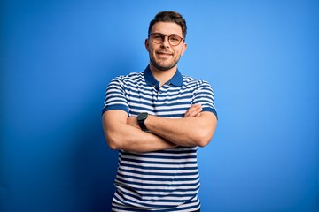 Young man with blue eyes wearing glasses and casual striped t-shirt over blue background happy face smiling with crossed arms looking at the camera. Positive person.