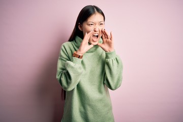 Young beautiful asian woman wearing green winter sweater over pink solated background Shouting angry out loud with hands over mouth