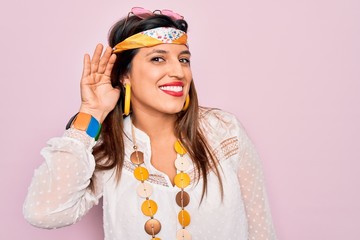 Young hispanic hippie woman wearing fashion boho style and sunglasses over pink background smiling with hand over ear listening an hearing to rumor or gossip. Deafness concept.