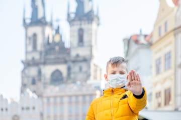 Fototapeta premium Boy wearing medical protective mask stands on the old town square in Prague and shows stop gesture. Coronavirus epidemic concept