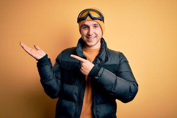 Young handsome caucasian man wearing hat, coat and ski glasses for winter and snow weather amazed and smiling to the camera while presenting with hand and pointing with finger.