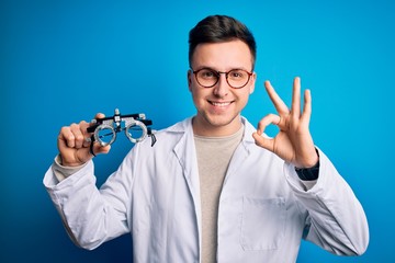Young handsome professional optic man holding optometrical glasses over blue background doing ok sign with fingers, excellent symbol
