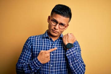 Young handsome latin man wearing casual shirt and glasses over yellow background In hurry pointing...