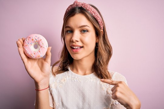 Young blonde girl eating sweet bakery sugar doughnut over pink isolated background with surprise face pointing finger to himself