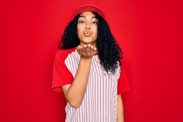 Young african american curly sportswoman wearing baseball cap and striped t-shirt looking at the camera blowing a kiss with hand on air being lovely and sexy. Love expression.