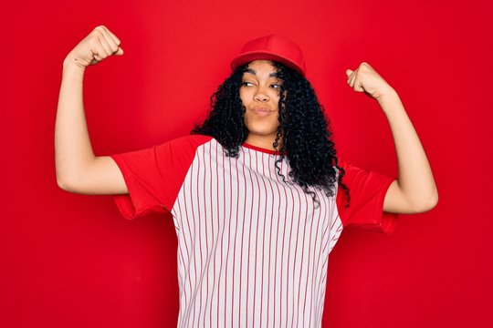 Young african american curly sportswoman wearing baseball cap and striped t-shirt showing arms muscles smiling proud. Fitness concept.
