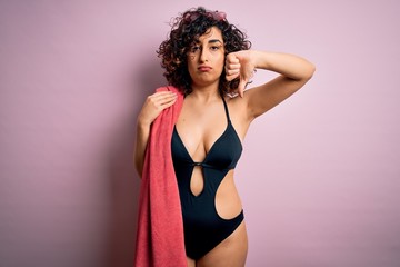 Obraz premium Young beautiful curly arab woman on vacation wearing swimsuit holding beach towel with angry face, negative sign showing dislike with thumbs down, rejection concept
