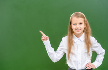 Smiling girl wearing eyeglasses points away on empty green chalkboard. Empty space for text