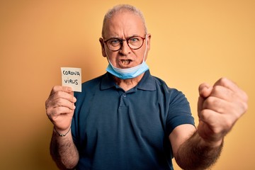 Middle age hoary man wearing medical mask holding reminder with coronavirus alert message annoyed and frustrated shouting with anger, crazy and yelling with raised hand, anger concept