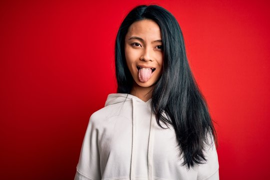 Young beautiful chinese sporty woman wearing sweatshirt over isolated red background sticking tongue out happy with funny expression. Emotion concept.