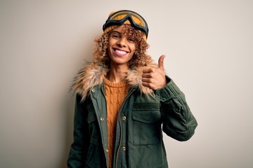 Young african american skier woman with curly hair wearing snow sportswear and ski goggles doing happy thumbs up gesture with hand. Approving expression looking at the camera showing success.