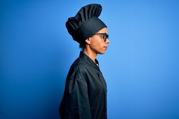 Young african american chef woman wearing cooker uniform and hat over blue background looking to side, relax profile pose with natural face and confident smile.