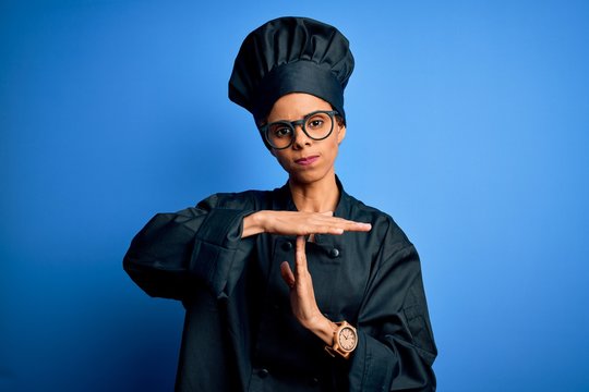 Young african american chef woman wearing cooker uniform and hat over blue background Doing time out gesture with hands, frustrated and serious face - Powered by Adobe