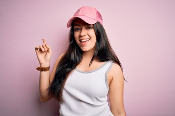 Young brunette woman wearing casual sport cap over pink background with a big smile on face, pointing with hand and finger to the side looking at the camera.