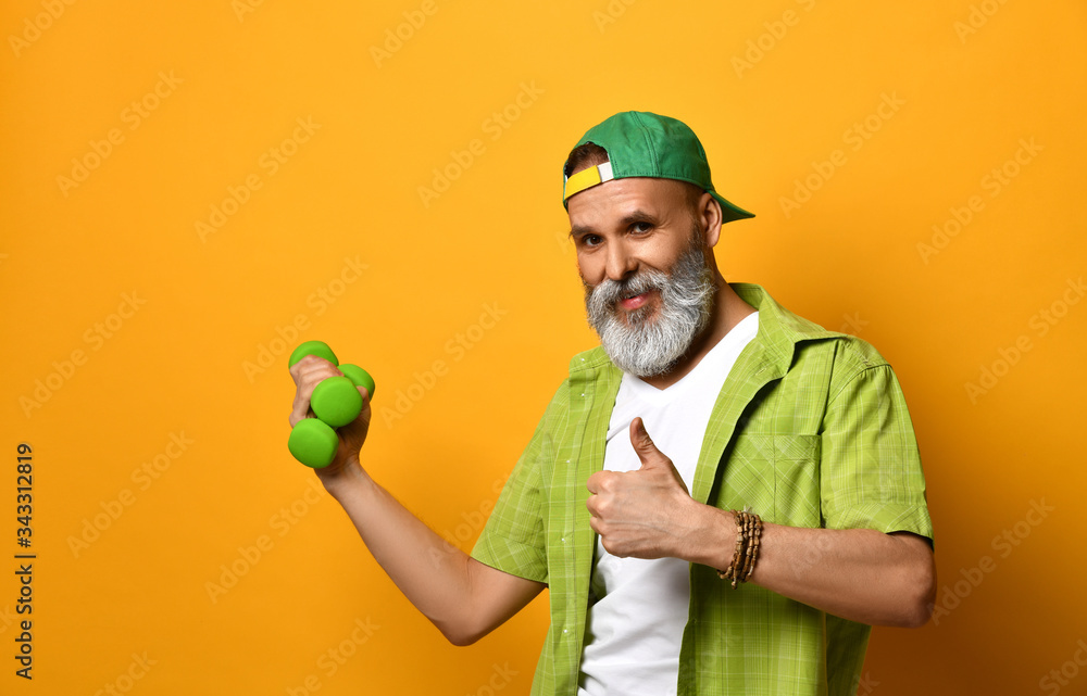 Wall mural grandpa in green cap and shirt, white t-shirt, bracelet. holding two dumbbells and pointing at them,