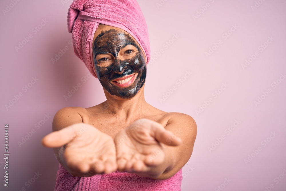 Wall mural middle age brunette woman wearing beauty black face mask over isolated pink background smiling with 