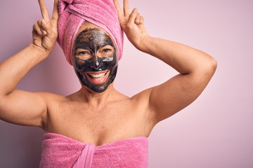 Middle age brunette woman wearing beauty black face mask over isolated pink background Posing funny and crazy with fingers on head as bunny ears, smiling cheerful